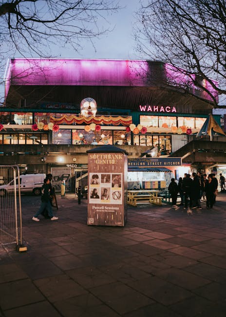 The image shows a lively outdoor area in front of a covered building, decorated with pink and red floral garlands and colorful banners beneath a large, illuminated, pink-lit roof structure. In the foreground, there are several people walking and standing, with some appearing to wait or engage in conversation. To the left, a chain-link fence partially encloses a small area, while a prominent wooden stand or sign displaying information about a documentary or event is positioned centrally, featuring black and white portraits and text. Behind the stand, a low terrace or platform area hosts additional seating and small food or drink stalls, including a sign for a Mexican street kitchen. Vehicles are parked further back near the building, which likely serves as a community or entertainment venue. Overhead, leafless trees extend their branches into a grey sky, suggesting either late autumn or winter. The scene captures a casual, social atmosphere consistent with a public gathering space, potentially associated with outdoor entertainment or festival activity, fitting within a context of alternative, private waste handling or rubbish clearance services offered by Rubbish Removal Waterloo.
