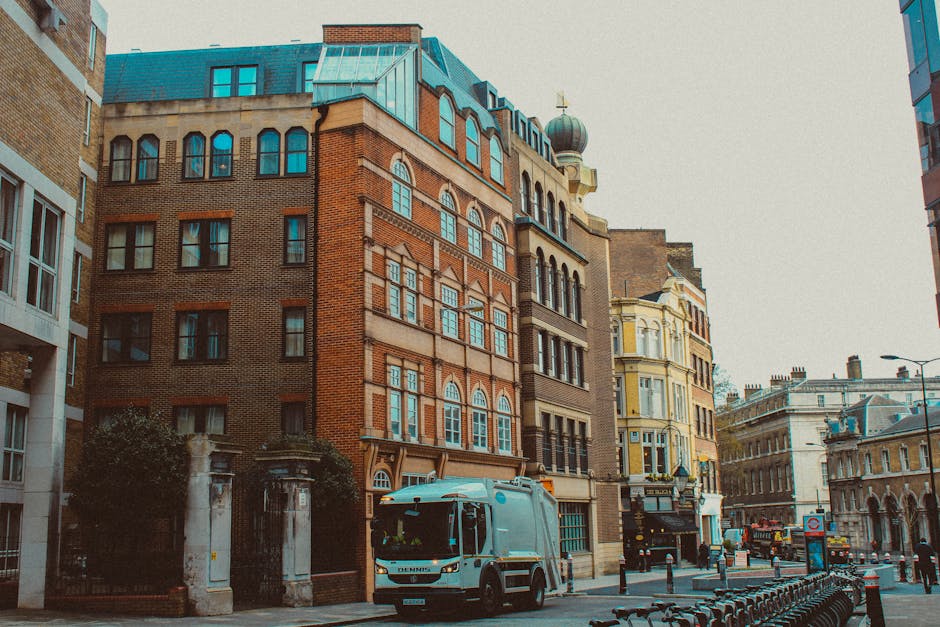 A street scene featuring a mixture of historical and modern buildings with distinct architectural details, including arched windows, ornate facades, and a dome-shaped turret on the top of one structure. The buildings are constructed from materials such as brick, stone, and plaster, with colors ranging from red brick to creamy yellow and light beige. In the foreground, a white and blue rubbish collection vehicle is parked on the pavement, positioned near a small section of sidewalk and some bollards that separate the pedestrian area from the road. To the left, a metal gate with stone pillars and greenery surrounds an entrance to a private property. The street appears quiet with minimal traffic, and the lighting suggests a daytime setting with overcast or diffused natural light. The scene subtly reflects an environment where private waste collection services, such as those provided by Rubbish Removal Waterloo, may operate independently of local authority rubbish collection, especially given the presence of a dedicated rubbish vehicle on the street.