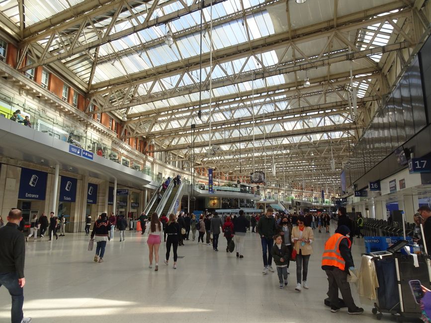 The image depicts the spacious interior of a busy railway station with a high, glass-paneled ceiling supported by metal beams, allowing natural light to flood the area. The station's main floor is populated with numerous travelers walking in various directions, some carrying luggage or backpacks. In the foreground, there is a person wearing an orange high-visibility vest attending to luggage near baggage trolleys, indicating an assistance or security role. The middle ground features a wide staircase and escalators leading to an upper level with additional seating and shops, while the background showcases ticket machines, information signs, and electronic displays. The station's walls are lined with large signage for entrances, ticketing, and platform numbers, with modern finishes and glass partitions. The environment appears bright and bustling, emphasizing the functional, organized space for passenger movement and transit. The scene relates to the broader context of transportation infrastructure, where waste management services like Rubbish Removal Waterloo may assist in maintaining cleanliness through alternative waste collection solutions amid high foot traffic.
